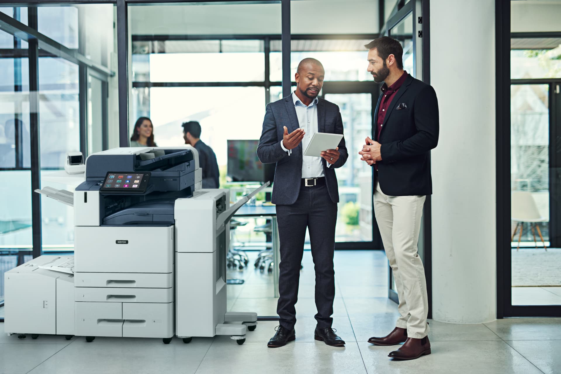 Xerox AltaLink C8200 series color copier with booklet maker finisher installed in a bright open-plan office while two colleagues review a job on a tablet